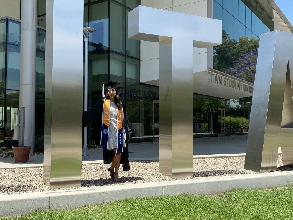 a woman in grad regalia stading outside near a large letter