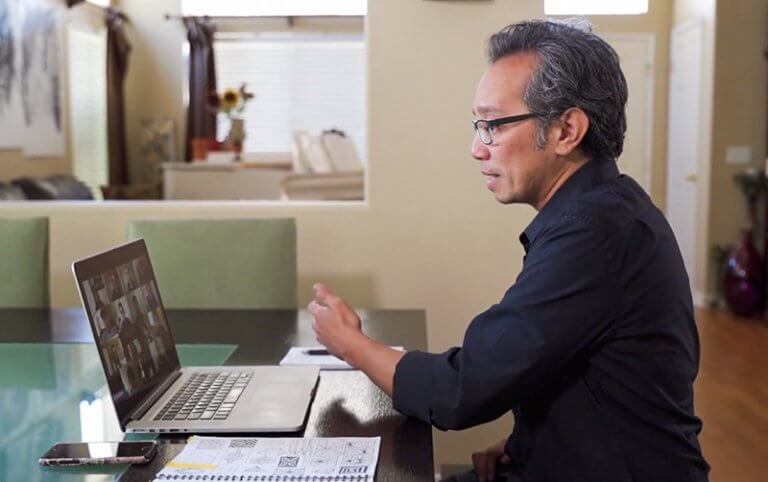 a man wearing eyeglasses sitting and looking at a laptop teaching an online class