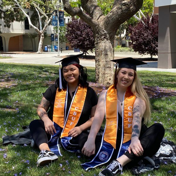 two women in grad regalia sitting on grass and smiling