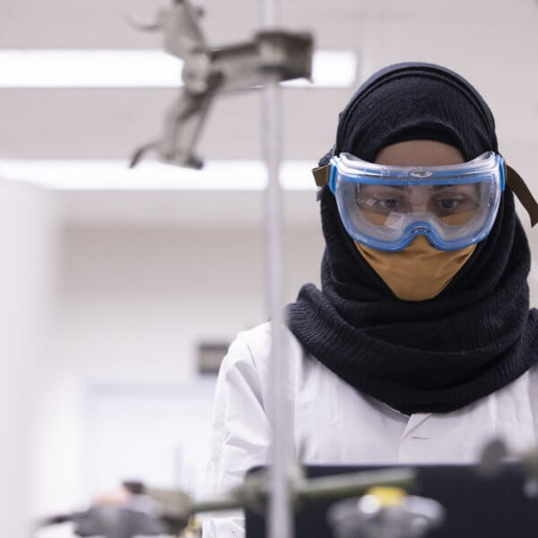 A young woman wearing a white lab coat, safety goggles and a face mask in a science lab